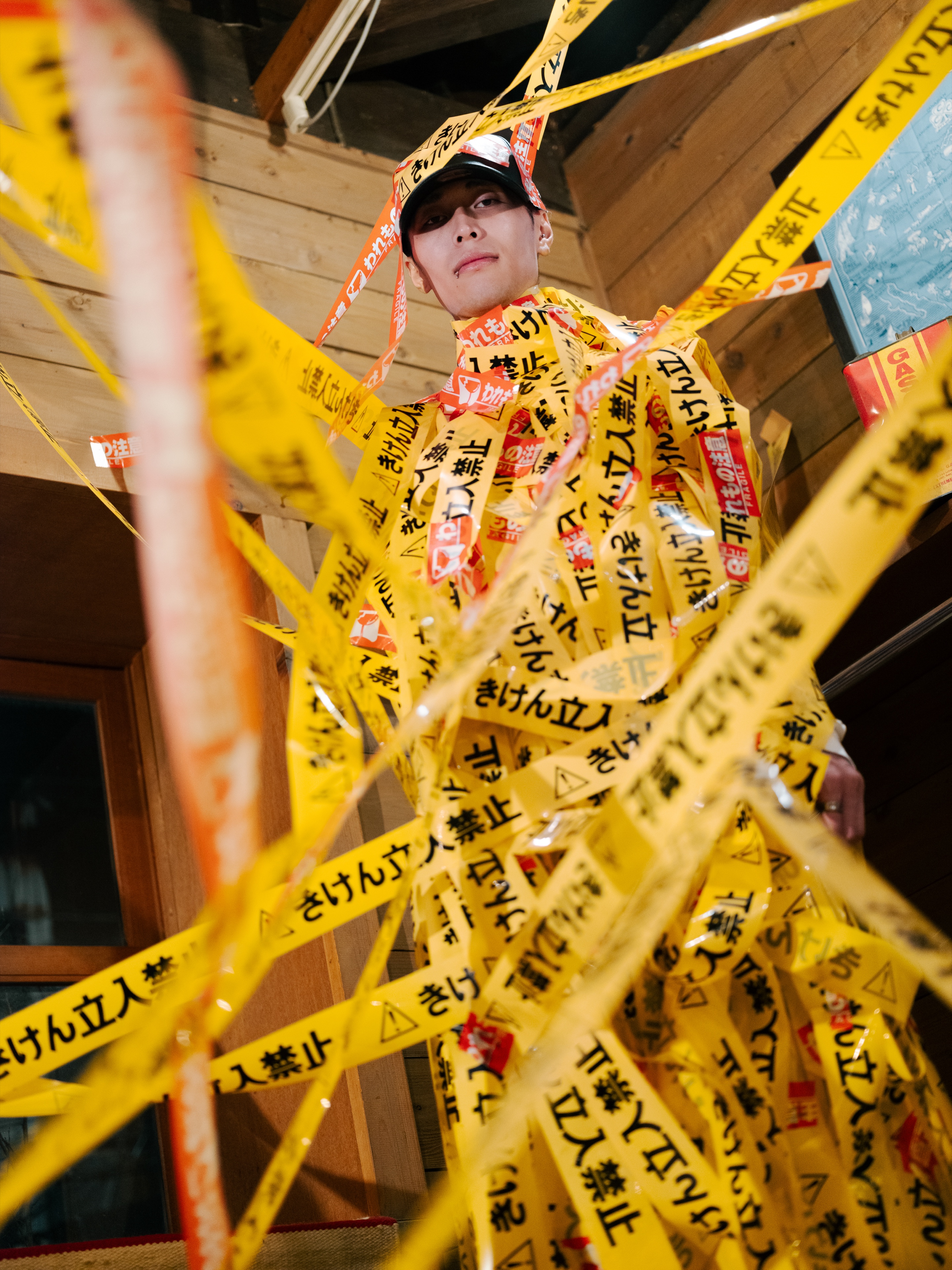 TAKAHASHI LAB ART — a person wrapped head to toe in yellow barrier tape, with more of the same tape stretched across the space around them